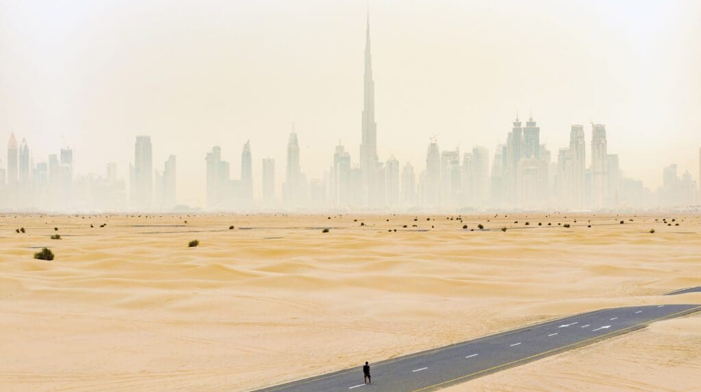 Aerial view of an unidentified person walking on a deserted road covered by sand dunes with the Dubai skyline in the background. Dubai, United Arab Emirates
