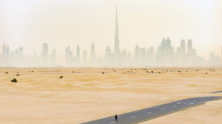 Aerial view of an unidentified person walking on a deserted road covered by sand dunes with the Dubai skyline in the background. Dubai, United Arab Emirates