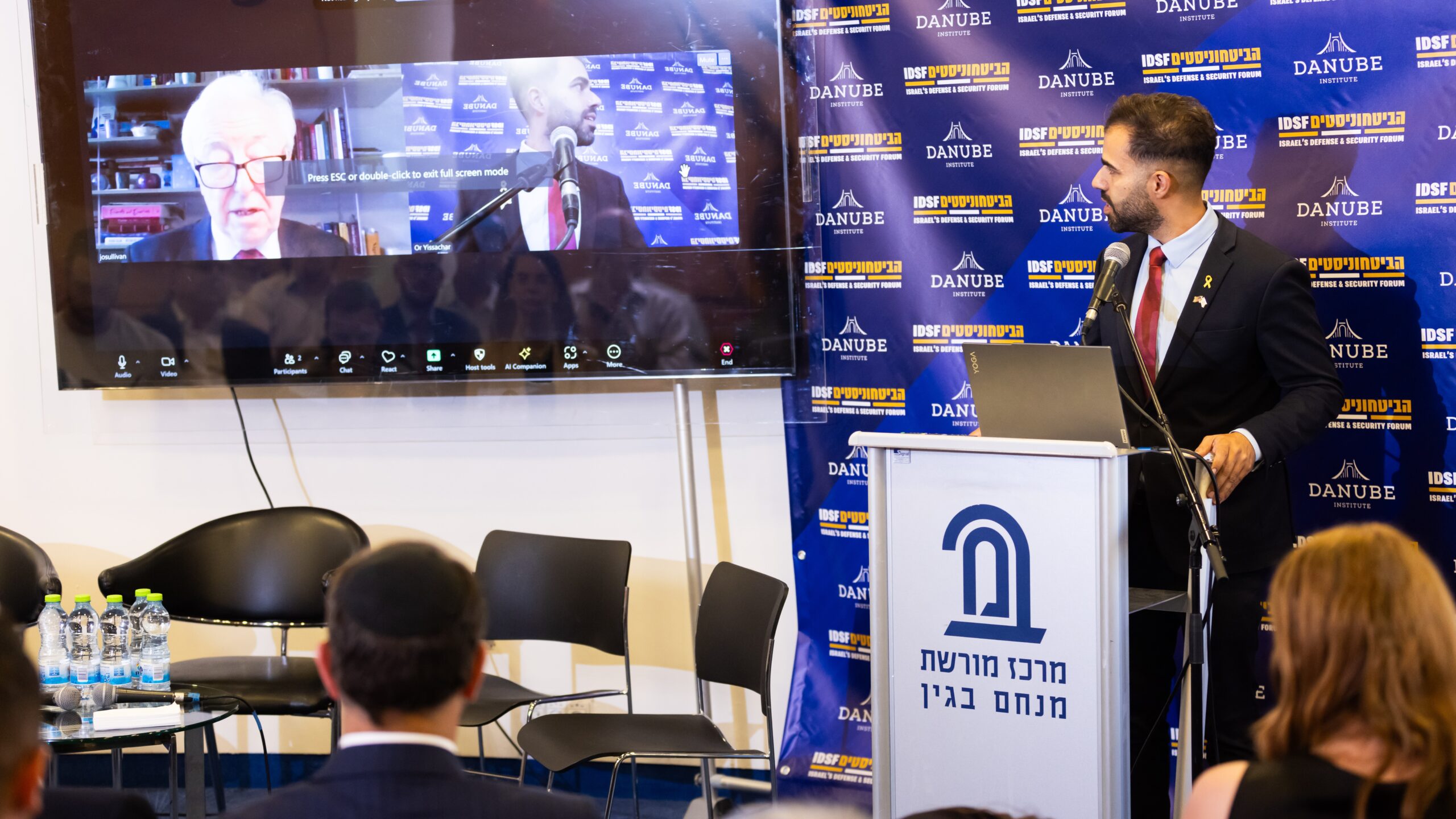 IDSF Research Department Head Or Yissachar (at the podium) and Danube Institute President John O’Sullivan (via video) greet the participants of the conference on 8 September 2024 in Jerusalem.